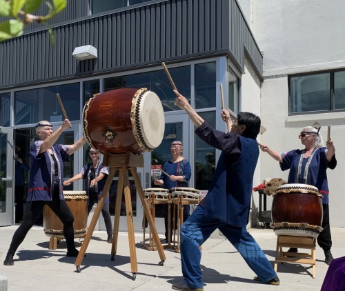 Two performers play a two-headed drum on an elevated stand with three other drummers in the background